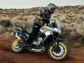 Hombre en moto china trail, recorriendo un camino polvoriento en un paisaje desértico con arbustos secos.