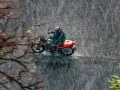 Un motociclista con casco y traje impermeable se aventura a conducir moto con lluvia intensa, rodeado de árboles y vegetación.
