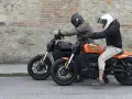 Dos personas en cascos montan dos motos custom electricas, una negra y otra naranja, frente a una pared de piedra.