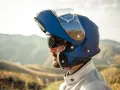 Hombre con barba y gafas de sol lleva un casco integral que se abre, azul, con visera levantada, listo para la aventura en la montaña.