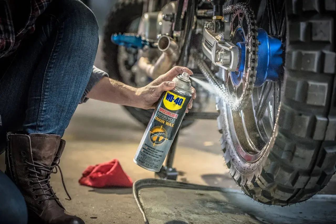 Persona en cuclillas aplica cera a la cadena de una moto. Mantenimiento esencial para engrasar cadena moto y asegurar un viaje suave.