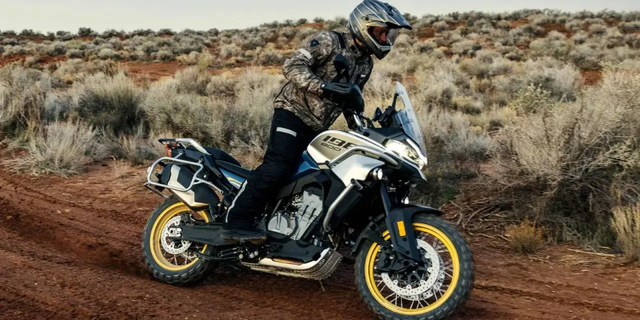 Hombre en moto china trail, recorriendo un camino polvoriento en un paisaje desértico con arbustos secos.