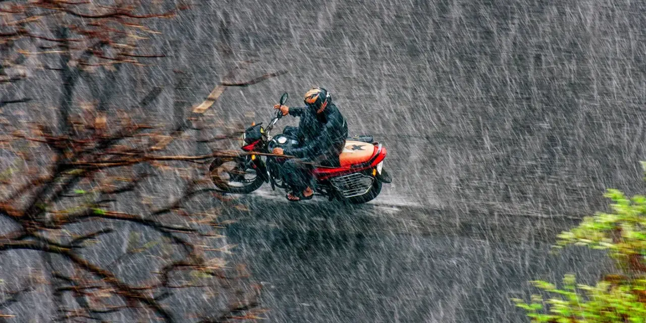 Un motociclista con casco y traje impermeable se aventura a conducir moto con lluvia intensa, rodeado de árboles y vegetación.