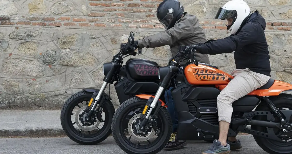 Dos personas en cascos montan dos motos custom electricas, una negra y otra naranja, frente a una pared de piedra.