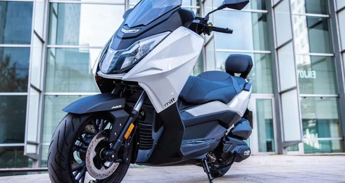 Scooter Mitt 330 GTS blanco y negro, con ABS, aparcado frente a un edificio moderno. ¡Sin problemas para disfrutar!