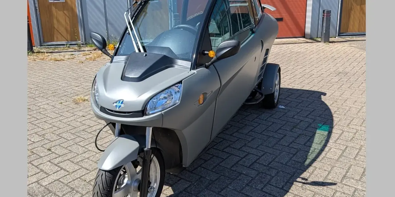 Un Carver One gris, un triciclo cerrado con techo panorámico, estacionado sobre adoquines.