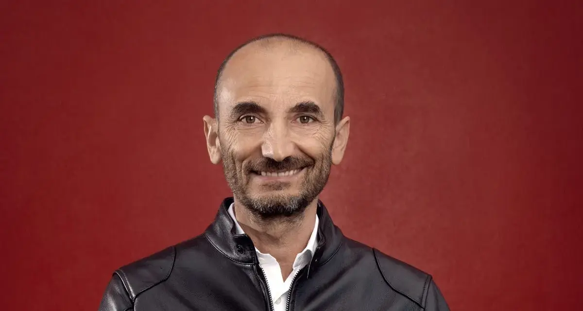 Claudio Domenicali, CEO de Ducati, sonríe con una chaqueta de cuero negra sobre una camisa blanca, sobre fondo rojo.