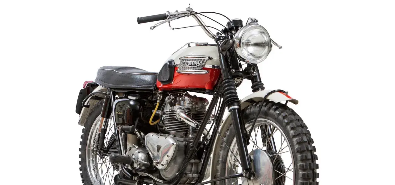 Clásica moto Triumph, modelo scrambler, con tanque bicolor rojo y blanco, motor expuesto y llantas todoterreno.