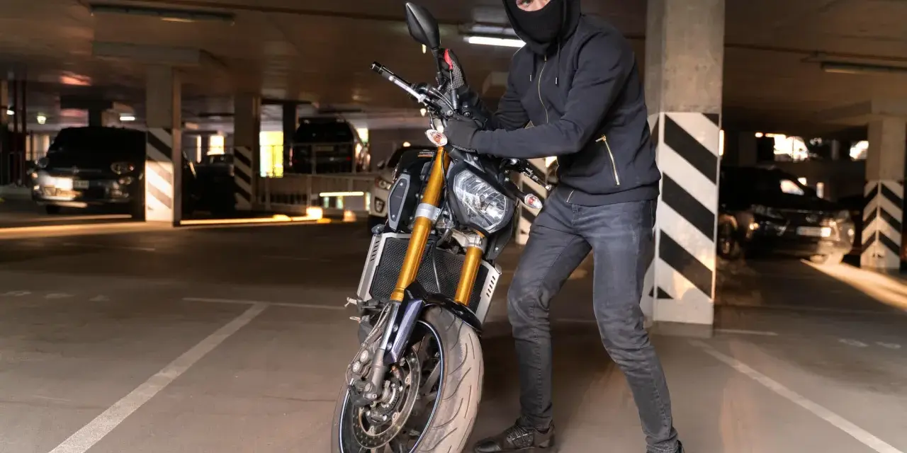 Ladrón con pasamontañas manipula una moto robada en un aparcamiento oscuro.