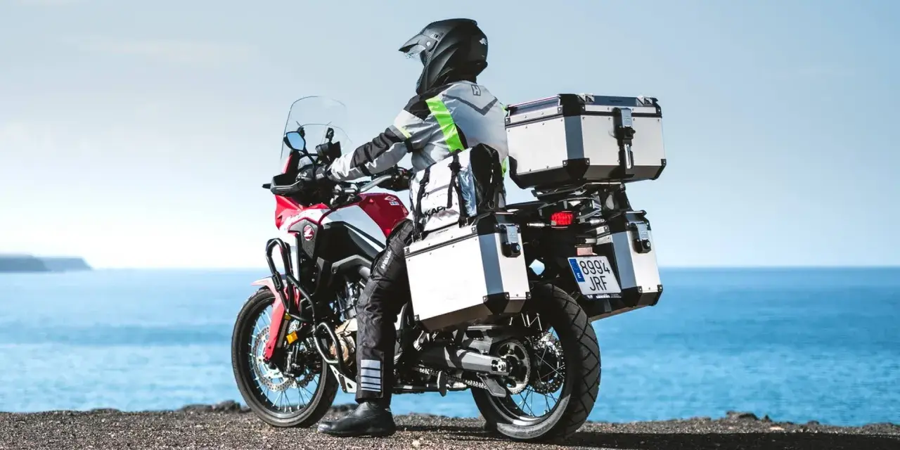 Motociclista con equipaje de **marca Kappa** listo para la aventura junto al mar.