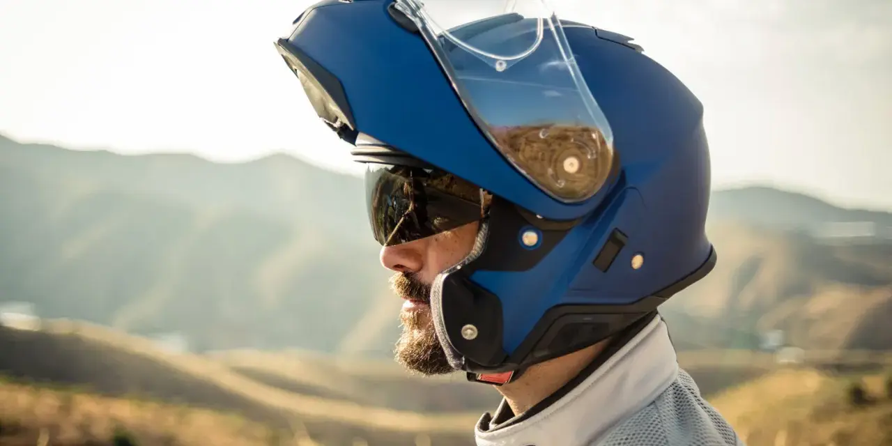 Hombre con barba y gafas de sol lleva un casco integral que se abre, azul, con visera levantada, listo para la aventura en la montaña.