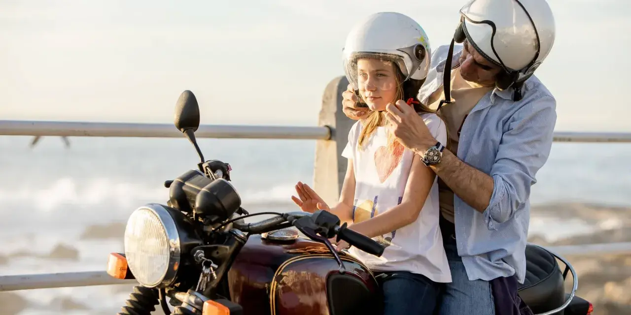 Un hombre ajusta el casco a un niño en una moto, listos para una aventura junto al mar.