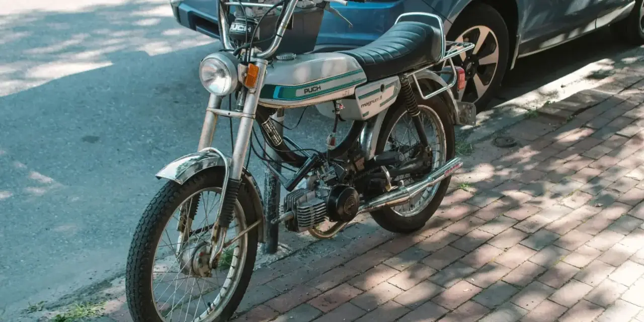 Ciclomotor antiguo Puch Magnum II plateado, listo para matricular. Aparcado junto a un coche azul en la acera.