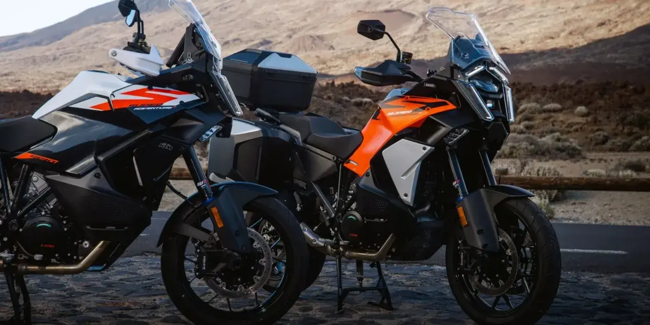 Dos motos KTM Super Adventure listas para la aventura. Modelos naranjas y blancos, con maletas, en un paisaje árido.