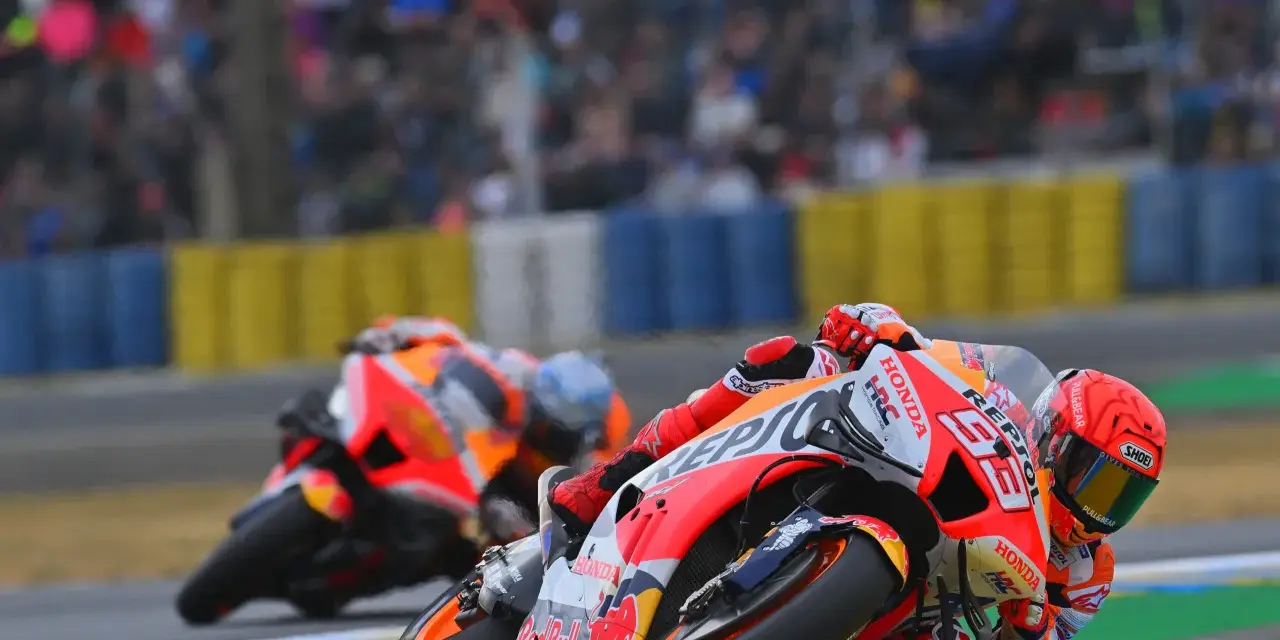 Dos pilotos de long lap motogp se inclinan en una curva, con el público borroso al fondo.