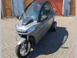 Un Carver One gris, un triciclo cerrado con techo panorámico, estacionado sobre adoquines.