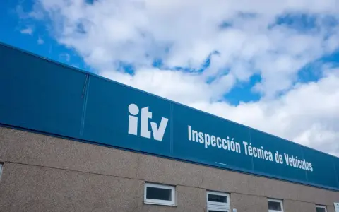 Edificio de la ITV bajo un cielo azul con nubes. El letrero blanco dice "itv Inspección Técnica de Vehículos".