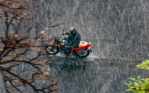 Un motociclista con casco y traje impermeable se aventura a conducir moto con lluvia intensa, rodeado de árboles y vegetación.