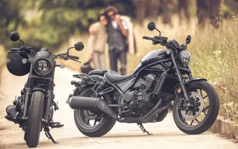 Dos motos negras, una con casco, listas para un viaje. Una pareja abraza en el fondo, disfrutando del paisaje. Una moto sin marchas, perfecta para la aventura.