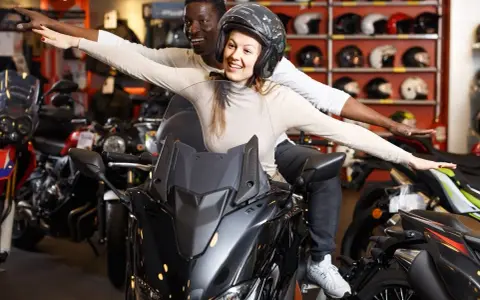 Pareja emocionada en una moto, soñando con su próxima aventura. La tienda de motos, con cascos en exhibición, parece el lugar perfecto para una reserva de dominio moto.