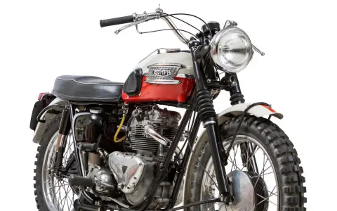 Clásica moto Triumph, modelo scrambler, con tanque bicolor rojo y blanco, motor expuesto y llantas todoterreno.