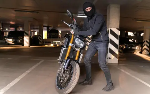 Ladrón con pasamontañas manipula una moto robada en un aparcamiento oscuro.