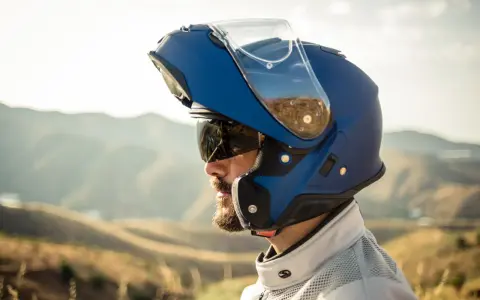 Hombre con barba y gafas de sol lleva un casco integral que se abre, azul, con visera levantada, listo para la aventura en la montaña.