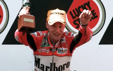 Ralf Waldmann, piloto de motos, celebra con un trofeo y un puño en alto, luciendo su traje de competición Marlboro.