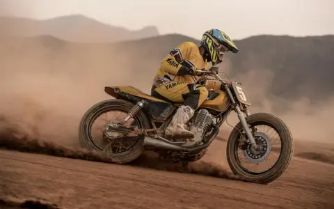 Piloto con casco y traje amarillo compite en una moto por una **dirt track**, levantando polvo en un paisaje desértico.