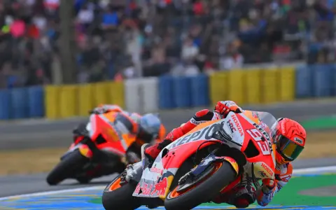 Dos pilotos de long lap motogp se inclinan en una curva, con el público borroso al fondo.