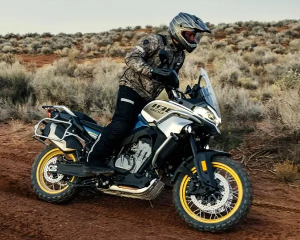 Hombre en moto china trail, recorriendo un camino polvoriento en un paisaje desértico con arbustos secos.