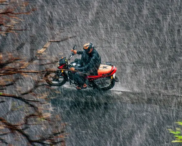 Un motociclista con casco y traje impermeable se aventura a conducir moto con lluvia intensa, rodeado de árboles y vegetación.