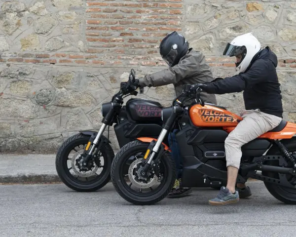 Dos personas en cascos montan dos motos custom electricas, una negra y otra naranja, frente a una pared de piedra.