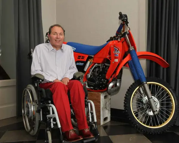 Andre Malherbe, leyenda del motocross, sonríe junto a su icónica moto Honda roja.