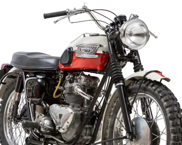 Clásica moto Triumph, modelo scrambler, con tanque bicolor rojo y blanco, motor expuesto y llantas todoterreno.