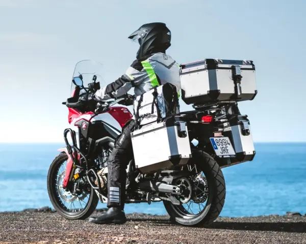 Motociclista con equipaje de **marca Kappa** listo para la aventura junto al mar.