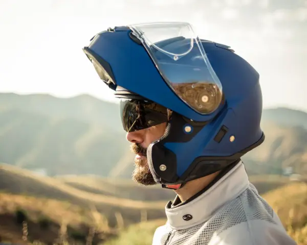 Hombre con barba y gafas de sol lleva un casco integral que se abre, azul, con visera levantada, listo para la aventura en la montaña.