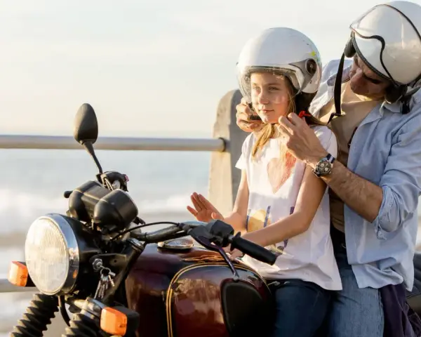 Un hombre ajusta el casco a un niño en una moto, listos para una aventura junto al mar.