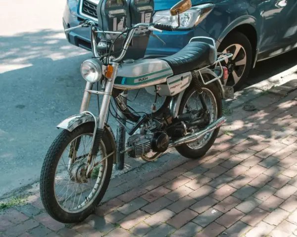 Ciclomotor antiguo Puch Magnum II plateado, listo para matricular. Aparcado junto a un coche azul en la acera.