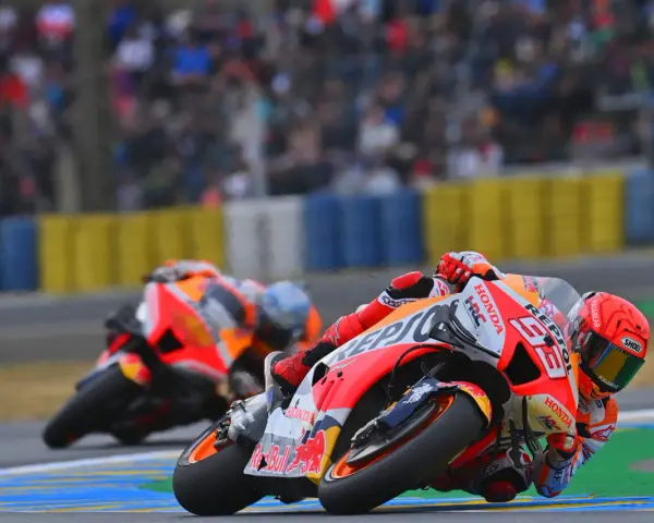 Dos pilotos de long lap motogp se inclinan en una curva, con el público borroso al fondo.