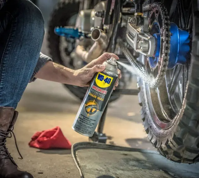 Persona en cuclillas aplica cera a la cadena de una moto. Mantenimiento esencial para engrasar cadena moto y asegurar un viaje suave.
