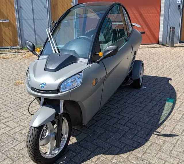 Un Carver One gris, un triciclo cerrado con techo panorámico, estacionado sobre adoquines.