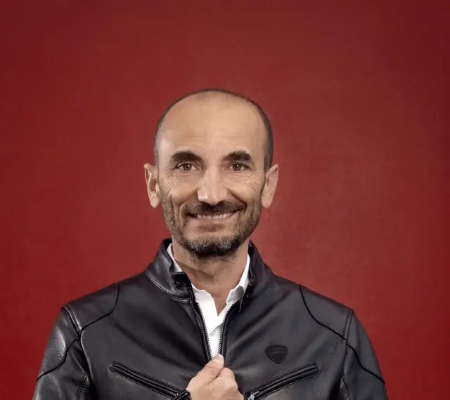 Claudio Domenicali, CEO de Ducati, sonríe con una chaqueta de cuero negra sobre una camisa blanca, sobre fondo rojo.