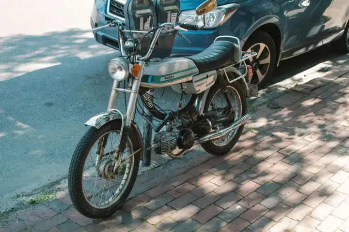 Ciclomotor antiguo Puch Magnum II plateado, listo para matricular. Aparcado junto a un coche azul en la acera.