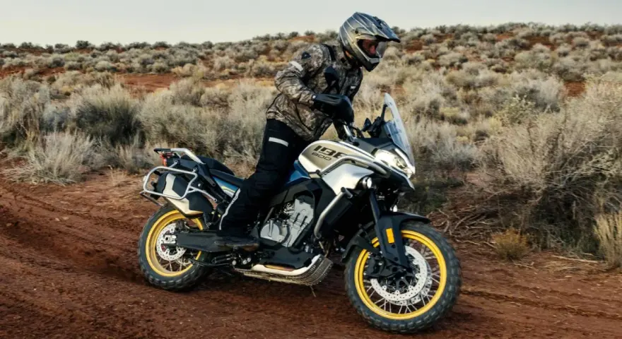 Hombre en moto china trail, recorriendo un camino polvoriento en un paisaje desértico con arbustos secos.