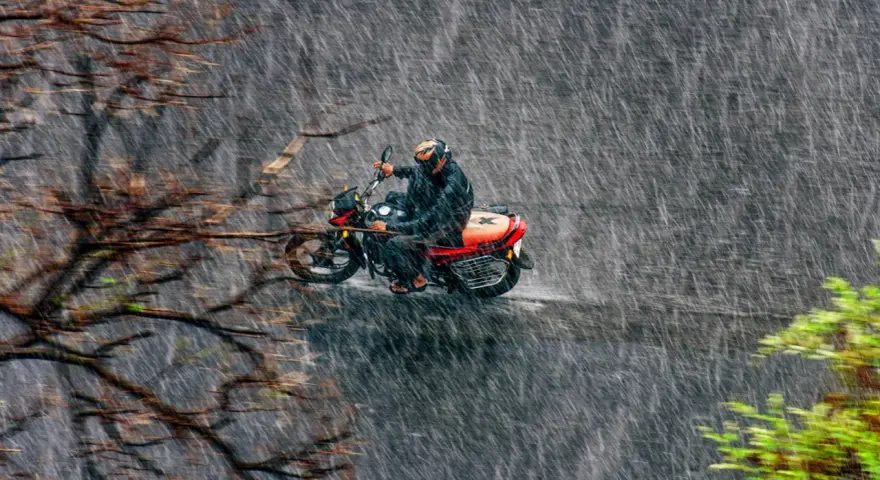 Un motociclista con casco y traje impermeable se aventura a conducir moto con lluvia intensa, rodeado de árboles y vegetación.