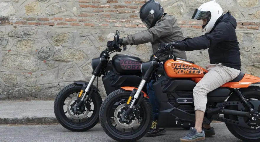 Dos personas en cascos montan dos motos custom electricas, una negra y otra naranja, frente a una pared de piedra.