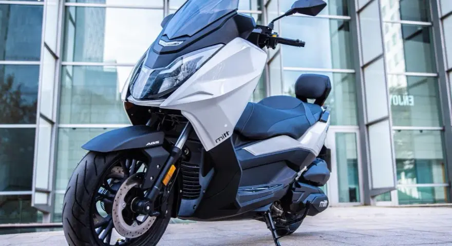 Scooter Mitt 330 GTS blanco y negro, con ABS, aparcado frente a un edificio moderno. ¡Sin problemas para disfrutar!
