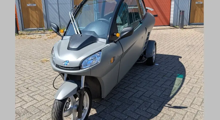 Un Carver One gris, un triciclo cerrado con techo panorámico, estacionado sobre adoquines.