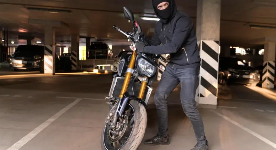 Ladrón con pasamontañas manipula una moto robada en un aparcamiento oscuro.